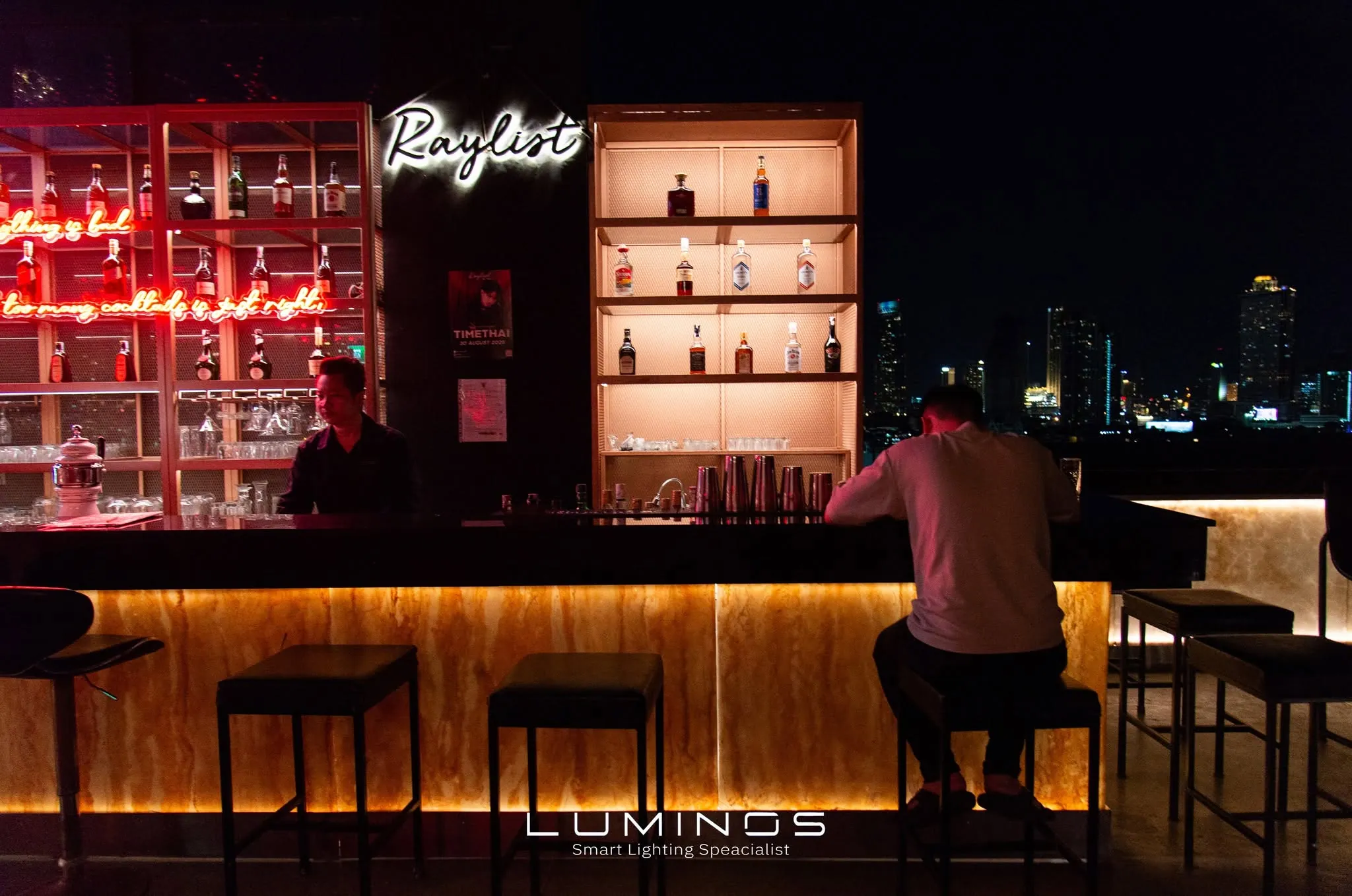 บาร์รูฟท็อปยามค่ำ ชั้นวางขวดเรืองแสงสีแดง ป้ายนีออน และเคาน์เตอร์หินอ่อนส่องไฟจากด้านใน