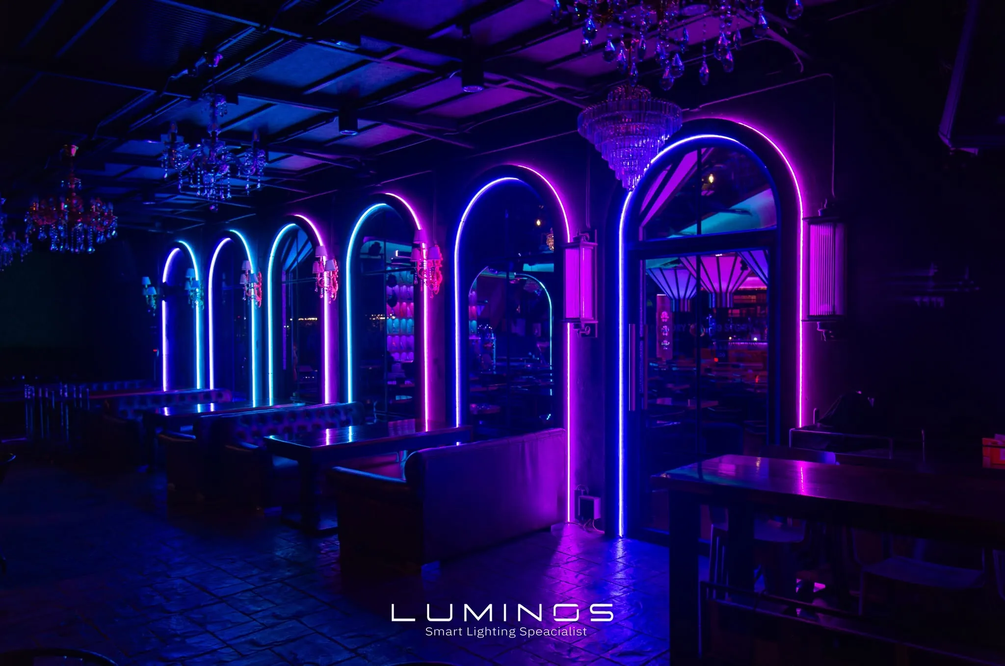 โซนที่นั่งริมทะเลยามค่ำ ที่นั่งโค้งอาบแสงสีส้มอุ่น ตัดกับโต๊ะที่ถูกไฟสีน้ำเงินและแดงส่องลงมา